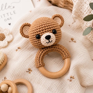 May include: A handmade crochet bear rattle with a wooden ring base. The bear is light brown with a cream-colored muzzle and black eyes. The rattle is on a cream-colored fabric with a floral pattern, alongside a wooden brush and a card that says "hello LITTLE ONE".
