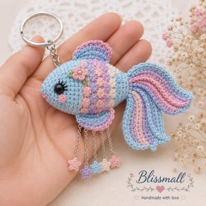 May include: A crocheted fish keychain featuring a light blue body with pink and purple scale details, a black eye, and a small flower accent. The fish has a flowing tail and five silver chains hanging from its underside, each ending in a small star charm.