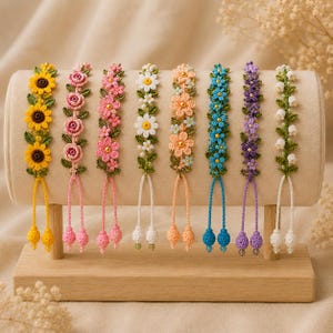 Könnte beinhalten: Acht Häkelarmbänder mit Blumenmotiven in Gelb, Rosa, Weiß, Pfirsich, Blau und Violett. Jedes Armband hat grüne Blätter, eine zentrale Perle und zwei herabhängende Kordelenden mit kleinen gehäkelten Kugeln, präsentiert auf einem zylindrischen Ständer.
