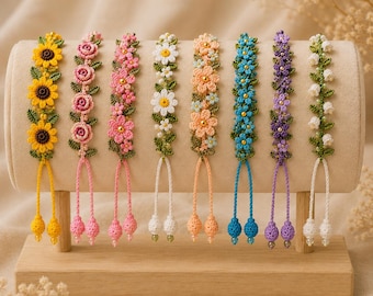May include: Eight crochet bracelets featuring floral designs in yellow, pink, white, peach, blue, and purple. Each bracelet has green leaves, a central bead, and two dangling cord ends with small crocheted spheres, displayed on a cylindrical stand.