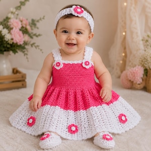 May include: A crochet baby dress featuring a bright pink bodice and a white skirt with pink flower accents. The dress has white straps with small flower details. A matching white crochet headband with a pink flower and white crochet booties.
