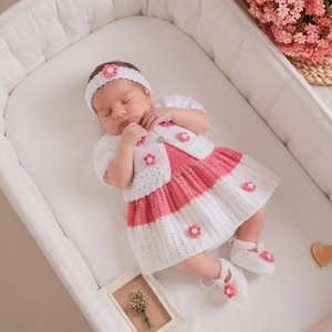 Può includere: Un completo per neonati lavorato all'uncinetto nei colori bianco e rosa, composto da un cardigan a maniche corte, una gonna plissettata, scarpine abbinate e una fascia. Ogni capo è decorato con piccole applicazioni floreali rosa e bianche.
