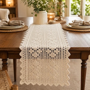 Puede incluir: Un camino de mesa rectangular de ganchillo en color blanco roto que presenta un intrincado patrón de rejilla calada y un borde festoneado. El camino está colocado sobre el centro de una mesa de comedor de madera, mostrando un tejido detallado.