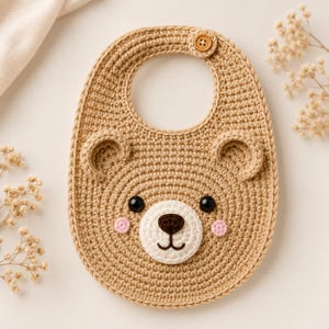 May include: A crocheted baby bib in a light brown color, shaped like a bear's face. The bib features black eyes, a white muzzle, a brown nose, and pink cheeks. A wooden button secures the bib at the neck.