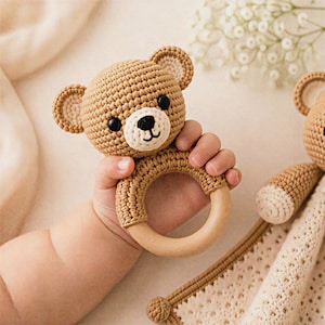 May include: A crocheted teddy bear rattle in shades of tan and cream, held by a baby's hand. The bear has black eyes and a white muzzle. The rattle features a wooden ring. A matching blanket is visible.