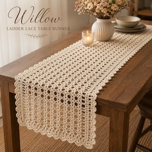 Pode incluir: Caminho de mesa creme com renda de escada sobre uma mesa de madeira. O caminho de mesa tem uma borda ondulada e padrões circulares abertos. O texto "Willow LADDER LACE TABLE RUNNER" está no canto superior esquerdo. Um vaso com flores, uma vela e uma tigela estão na mesa.