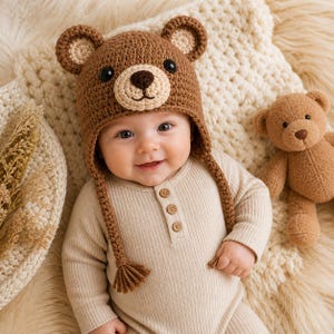 May include: A baby wearing a brown crochet bear hat with ear flaps and a matching teddy bear. The baby is dressed in a beige ribbed onesie with wooden buttons, lying on a cream-coloured blanket.