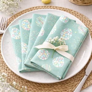 May include: Four folded teal napkins with a pastel egg design, tied with a cream ribbon and a sprig of white flowers, on a white plate. A knife and fork are on either side. A basket of Easter eggs is in the background.