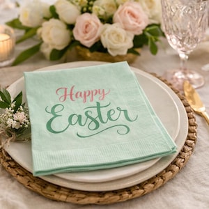 Può includere: Un'apparecchiatura pasquale festosa con un tovagliolo verde chiaro con la scritta "Happy Easter" in rosa e verde. La tavola è apparecchiata con posate dorate, piatti bianchi e una composizione floreale.