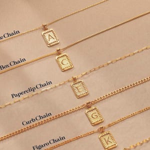 May include: Five gold-toned necklaces with rectangular pendants featuring letters A, C, E, G, and K. The chains are labeled as Cable Chain, Box Chain, Paperclip Chain, Curb Chain, and Figaro Chain, each showcasing a distinct link style and design.