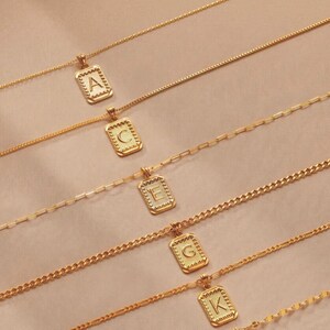 May include: Five gold-toned necklaces, each featuring a rectangular pendant with a scalloped edge and a single embossed letter. The letters A, C, E, G, and K are displayed on the pendants. Each necklace has a distinct chain style, including cable, link, and curb chain designs.