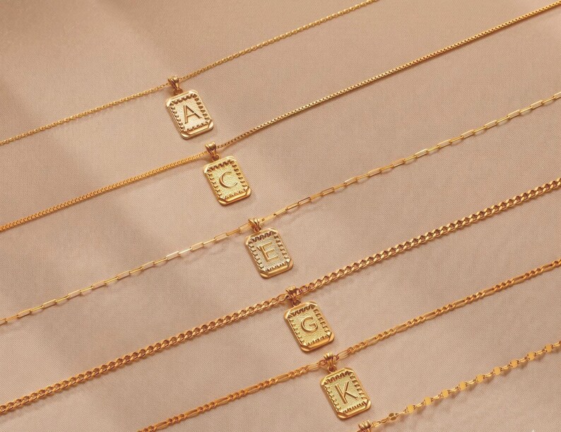 May include: Five gold-toned necklaces, each featuring a rectangular pendant with a scalloped edge and a single embossed letter. The letters A, C, E, G, and K are displayed on the pendants. Each necklace has a distinct chain style, including cable, link, and curb chain designs.