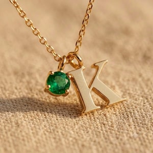 May include: A gold-colored necklace with a pendant featuring the letter "K" and a round, emerald-green gemstone. The necklace chain and pendant are made of a shiny, gold-toned metal. The gemstone is set in a gold-colored bezel.