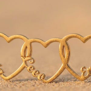 May include: A gold-toned necklace featuring three interconnected heart-shaped pendants. Each heart is engraved with a name in a cursive script: Aria on the left, Leea in the center, and Emma on the right. The piece includes a delicate chain.