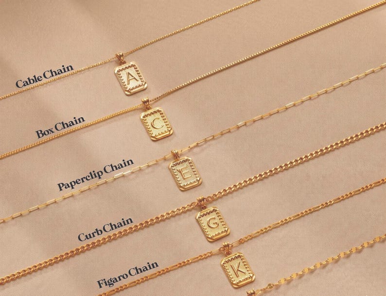 May include: Five gold-toned necklaces with rectangular pendants featuring letters A, C, E, G, and K. The chains are labeled as Cable Chain, Box Chain, Paperclip Chain, Curb Chain, and Figaro Chain, each showcasing a distinct link style and design.