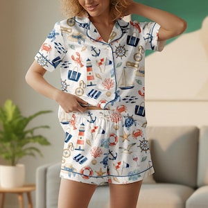 May include: White pajama set with a nautical theme. The short-sleeved top and shorts feature a pattern of anchors, sailboats, lighthouses, and marine life in blue, red, and beige. The set has blue trim.