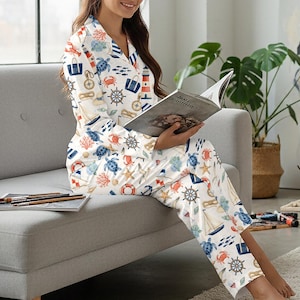 May include: White pajama set with a nautical theme, featuring illustrations of sailboats, turtles, anchors, and lighthouses in blue, red, and beige. The long-sleeved top has a collared neckline and button closure. The pants have a relaxed fit.