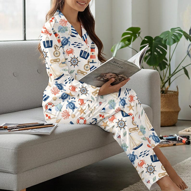 May include: White pajama set with a nautical theme, featuring illustrations of sailboats, turtles, anchors, and lighthouses in blue, red, and beige. The long-sleeved top has a collared neckline and button closure. The pants have a relaxed fit.