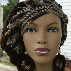 May include: A brown and beige crocheted hat with a unique design. The hat is worn on a mannequin with a neutral expression. The hat has a loose, flowing style and is made with a variety of stitches.