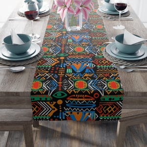 May include: A black table runner with a vibrant, geometric pattern in orange, blue, green, and white. The design features tribal-inspired shapes and symbols. The runner is placed on a wooden table set for a meal, with plates, glasses, and flowers.