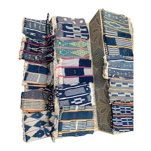 May include: A display of folded, patterned textiles in shades of blue, white, pink, and yellow. The fabrics feature geometric and striped designs, with fringed edges. The textiles are arranged in a multi-tiered, decorative display.