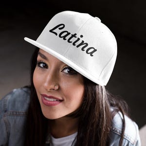 Może przedstawiać: Biała czapka z daszkiem z napisem "Latina" w czarnym skrypcie. Czapka ma płaski daszek. Osoba nosząca czapkę ma również na sobie dżinsową kurtkę.