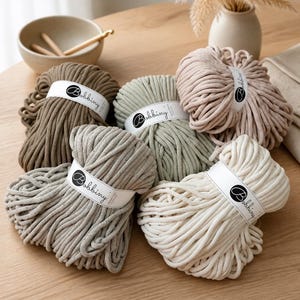 May include: Five skeins of thick, braided cotton cord in shades of beige, sage green, light brown, and off-white. Each skein features a white paper label with the black text Bobbiny. The cord has a smooth, tubular texture suitable for crafting.