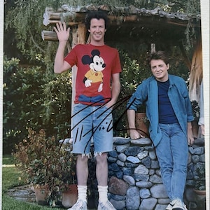 May include: A photograph featuring two men posing outdoors. One man wears a red t-shirt with a Mickey Mouse graphic, denim shorts, and white sneakers. The other man wears a blue shirt and jeans. The image is signed.