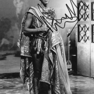 May include: Black and white photograph of a person in a classical costume. The person is wearing a tunic, cape, and sandals. The image has an autograph in the upper right corner.