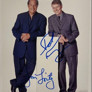May include: A photograph of two men in suits posing together. One man wears a navy blue suit, and the other wears a gray suit with a patterned tie. The photo includes blue signatures on a white surface.