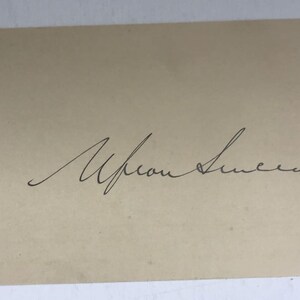 May include: A beige card with a handwritten signature in black ink. The signature appears to read "Upson Sinclair" in cursive script. The card has a slightly aged appearance.