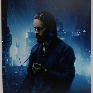 May include: A signed photograph featuring a man in a dark blue robe, set against a futuristic cityscape backdrop with rain. The image has a blue and gray color scheme, with a signature in blue ink.