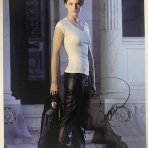 May include: A signed photograph of a woman wearing a white V-neck top and black leather trousers. She is holding a black jacket and standing in front of a building with columns. The photograph includes an autograph in black ink.