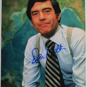 Pode incluir: Fotografia assinada de um homem usando uma camisa azul clara, gravata listrada e suspensórios. O fundo apresenta um mapa. A assinatura é em tinta azul.