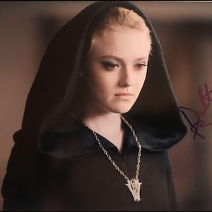Puede incluir: Retrato de una persona de piel clara, cabello rubio y ojos rojos, con una capa con capucha negra y un collar plateado. La imagen tiene una firma en la esquina superior derecha.