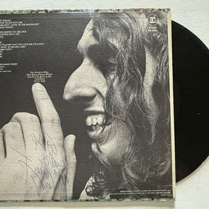 Può includere: Album in vinile vintage con un'immagine in bianco e nero del volto e della mano di una persona, con la firma dell'artista. La copertina dell'album include i titoli delle canzoni e le informazioni sull'artista. È visibile anche un disco in vinile nero.