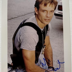 May include: A photograph of a man with short, light brown hair, wearing a gray t-shirt, blue jeans, and a black vest. He has a badge on a chain around his neck and a signature in blue ink. The background is blurred.