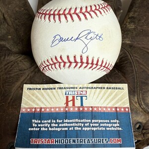 May include: A white baseball with red stitching and a blue autograph, resting on a brown leather baseball glove. A card with the text "TRISTAR HIDDEN TREASURES AUTOGRAPHED BASEBALL" is in front of the glove.
