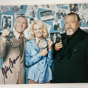 May include: A vintage photograph of three people holding microphones. The man on the left wears a tan suit and holds a microphone with "NBC" on it. The woman in the centre wears a blue blouse. The man on the right wears a black suit.