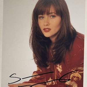 May include: A portrait of a woman with long, layered brown hair and bangs. She wears a red velvet top with gold embroidery and a signature in black ink. The background is a plain, light gray.