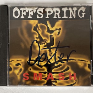 May include: A compact disc case featuring the band name "OFFSPRING" in white block letters above the word "SMASH" in red. The cover art displays a skeleton against a yellow and brown background. The signature "Dexter" is written in blue ink.