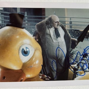 Może przedstawiać: Zdjęcie przedstawiające postać z filmu obok dużej, żółtej konstrukcji w kształcie kaczki. Postać ma na sobie garnitur i trzyma parasol. Obraz jest podpisany niebieskim atramentem. Tło sugeruje plan filmowy.