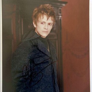May include: A signed photograph of a person with reddish-brown hair and pale skin, wearing a dark grey coat over a black polo neck. The background features a dark wood cabinet and a reddish-brown wall.