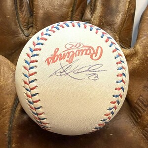 Może przedstawiać: Biała piłka baseballowa Rawlings z czerwonymi i niebieskimi szwami, posiadająca podpis czarnym tuszem na powierzchni. Piłka jest umieszczona na tle zużytej, brązowej skórzanej rękawicy baseballowej o widocznej fakturze.