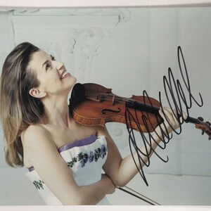May include: A photograph of a woman holding a violin. The violin is brown with black details. The woman is wearing a white dress with purple trim and floral details. The image is signed in black ink.