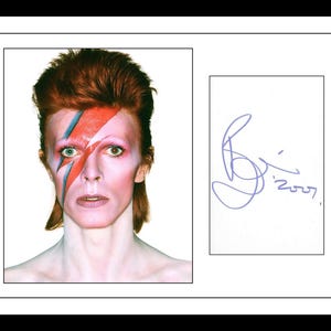 Puede incluir: Un retrato enmarcado de David Bowie con su icónico maquillaje de rayo rojo y azul, luciendo un peinado mullet de color castaño rojizo. Junto al retrato hay una tarjeta blanca con una firma en tinta morada y el año 2007.