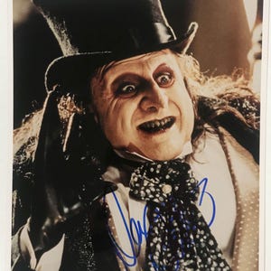 May include: A color photograph of a man in a top hat and formal attire, with a sinister expression. He has pale skin, dark eye makeup, and a black and white polka-dot scarf. The image is signed in blue ink.