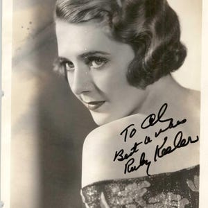 May include: A vintage black and white photograph of a woman with dark, wavy hair, looking over her shoulder. The photo includes an autograph that reads "To Al Best Wishes Ruby Keeler."