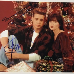 Könnte beinhalten: Ein Foto mit einem Mann und einer Frau, die vor einem Weihnachtsbaum posieren. Der Mann trägt ein dunkelrotes kariertes Hemd über einem weißen Hemd und einer blauen Jeans. Die Frau trägt ein dunkelrotes Oberteil. Beide haben Autogramme auf dem Foto.