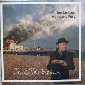 May include: Album cover for "Hope and Fury" by Joe Jackson. The image features a man in a hat holding a teacup on a beach with a pier in the background. The pier is partially on fire. The album title and artist name are visible.